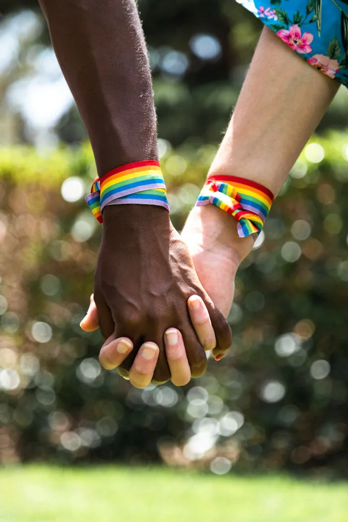 Two people holding hands outdoors, each wearing a rainbow Pride wristband
