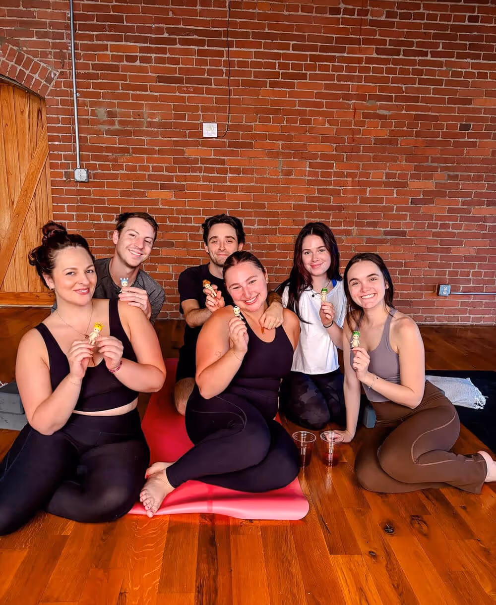 Group of six young adults sitting on a wooden floor with yoga mats, smiling and holding small figurines against a brick wall background.