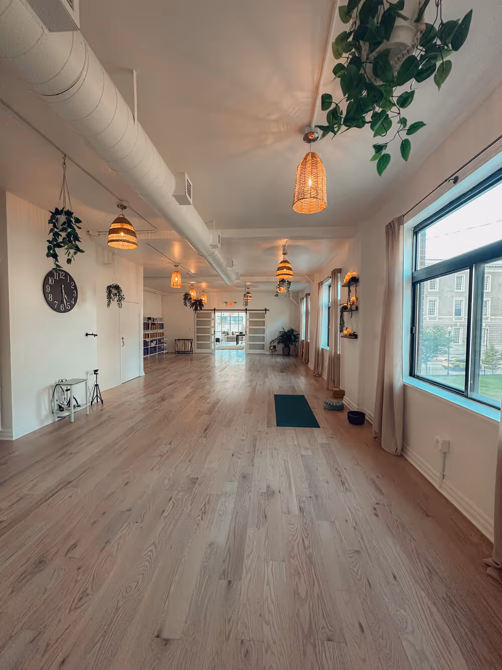 Spacious yoga studio with wooden floors, hanging plants, wicker pendant lights, large windows, and a yoga mat near the right wall.