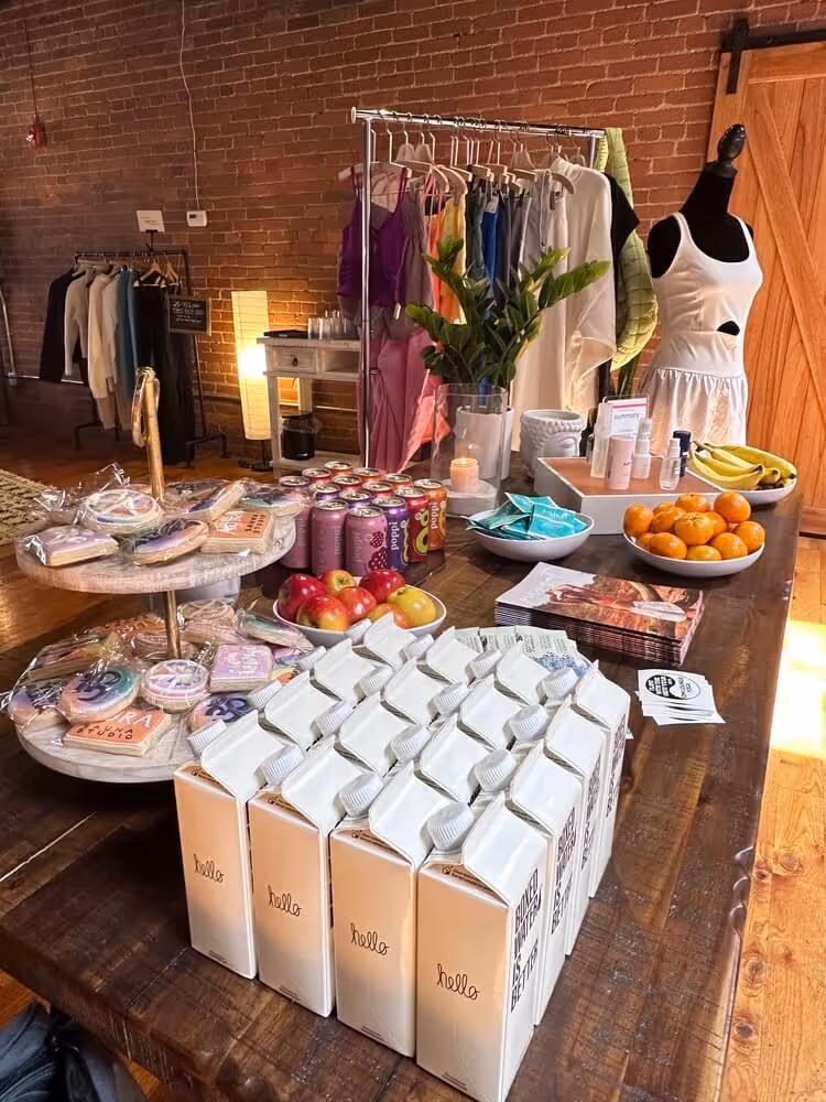 Wooden table displaying cartons of oat milk, bowls with apples, bananas, and oranges, canned drinks, packaged cookies, skincare products, and a clothing rack with colorful garments in the background.