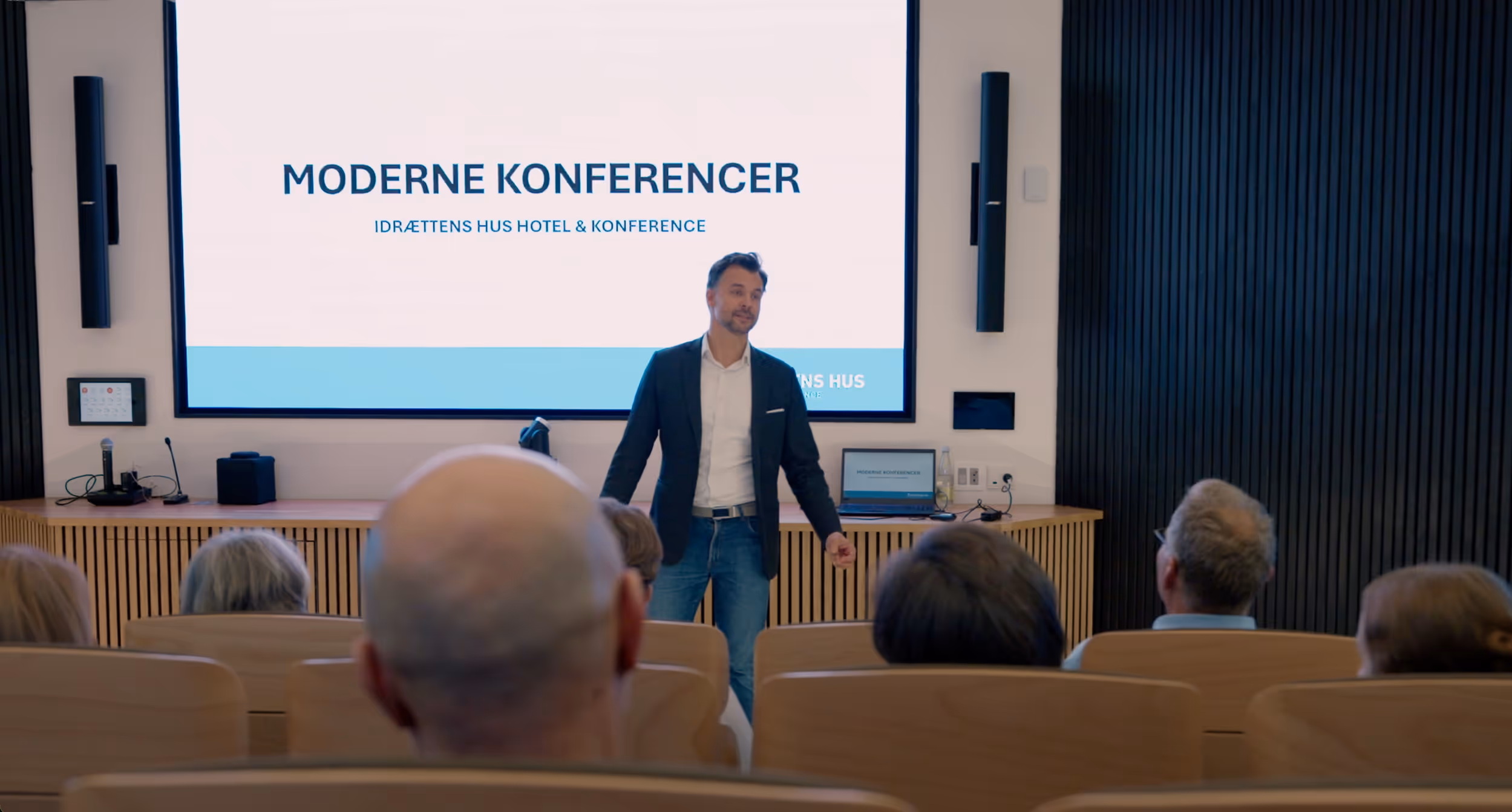 Mand holder præsentation foran et publikum i et moderne konferencelokale med stort skærm bag sig.