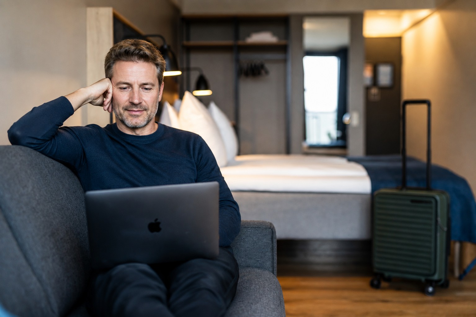 Hotel room. A woman is sitting on a couch with her laptop. Her suitcase is standing next to the double bed in the background.