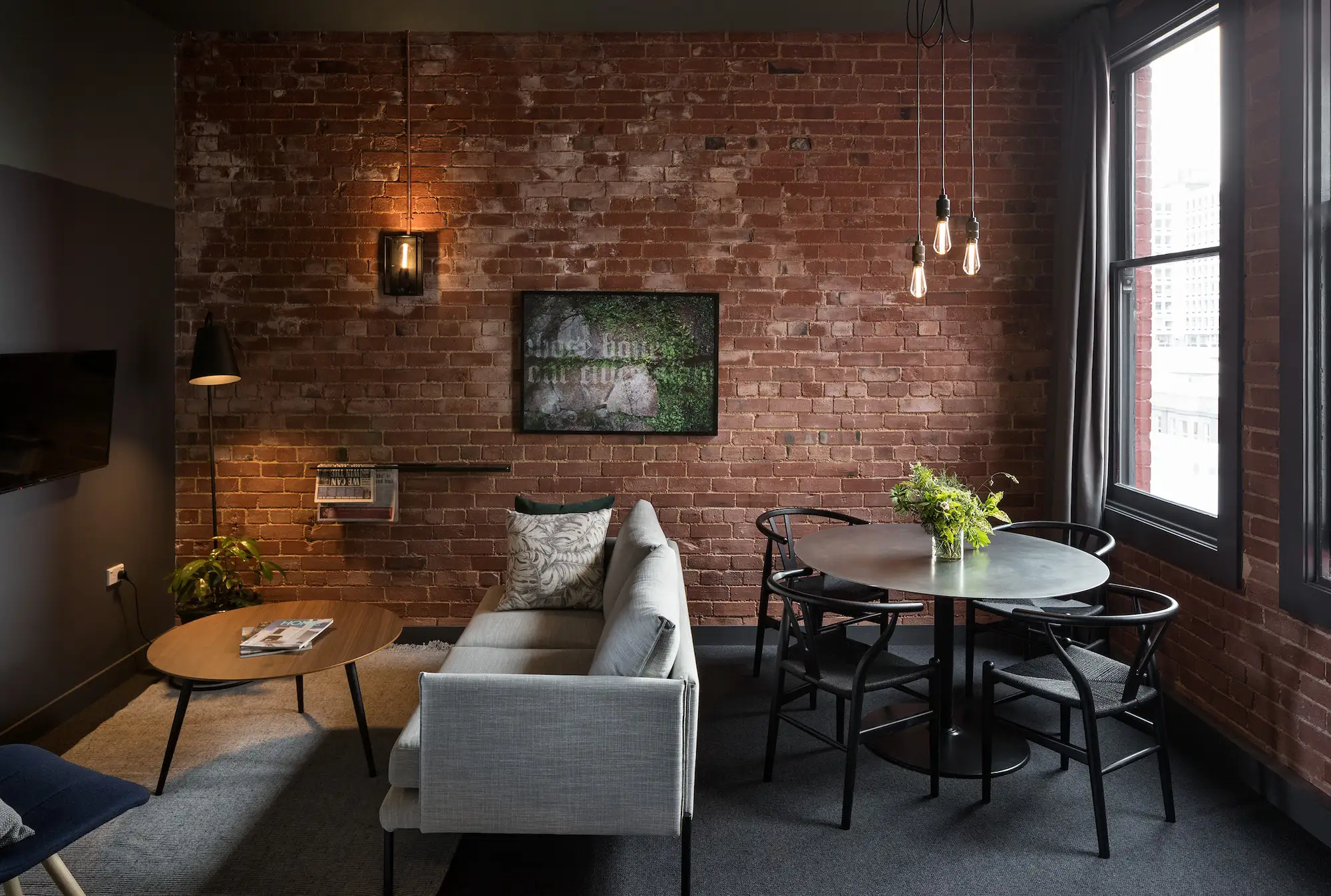 Modern living room with brick wall, grey sofa, round wooden coffee table, black dining table with four chairs, and large windows.