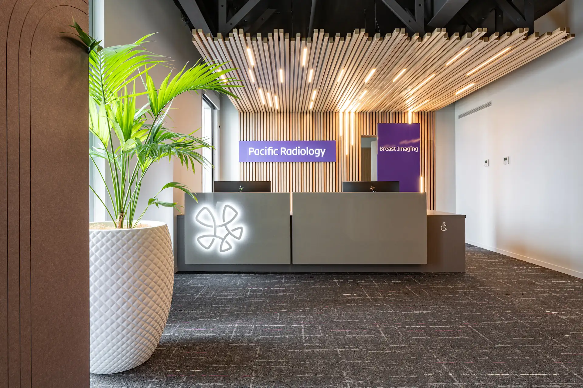 Reception area of Pacific Radiology with modern desk, illuminated logo, wooden slat ceiling, and large green plant in white pot.