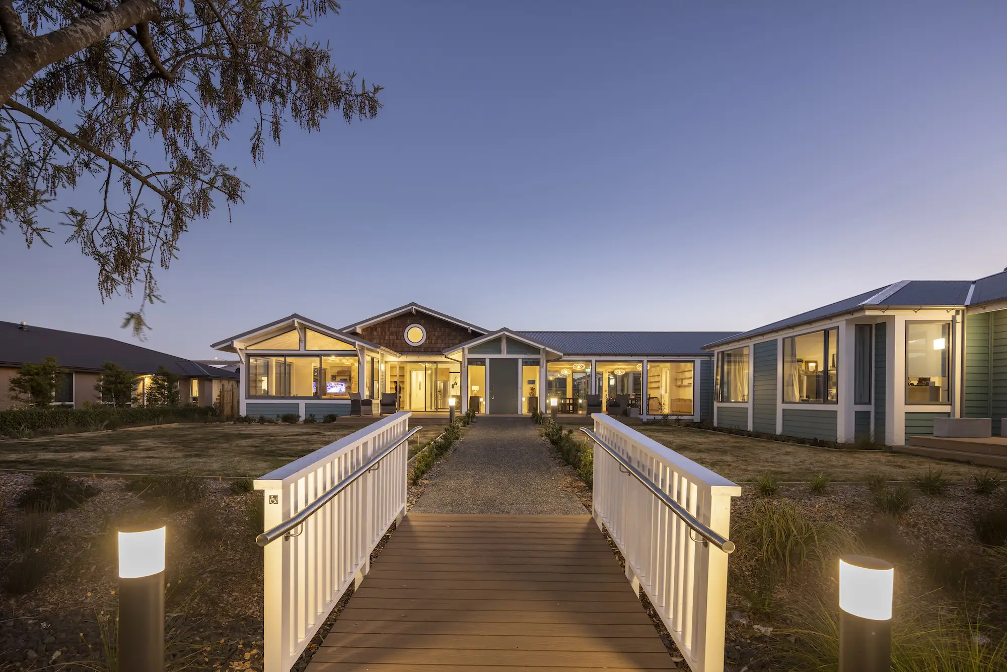 Modern single-story house with illuminated large windows and a ramped wooden walkway leading to the front door at dusk.