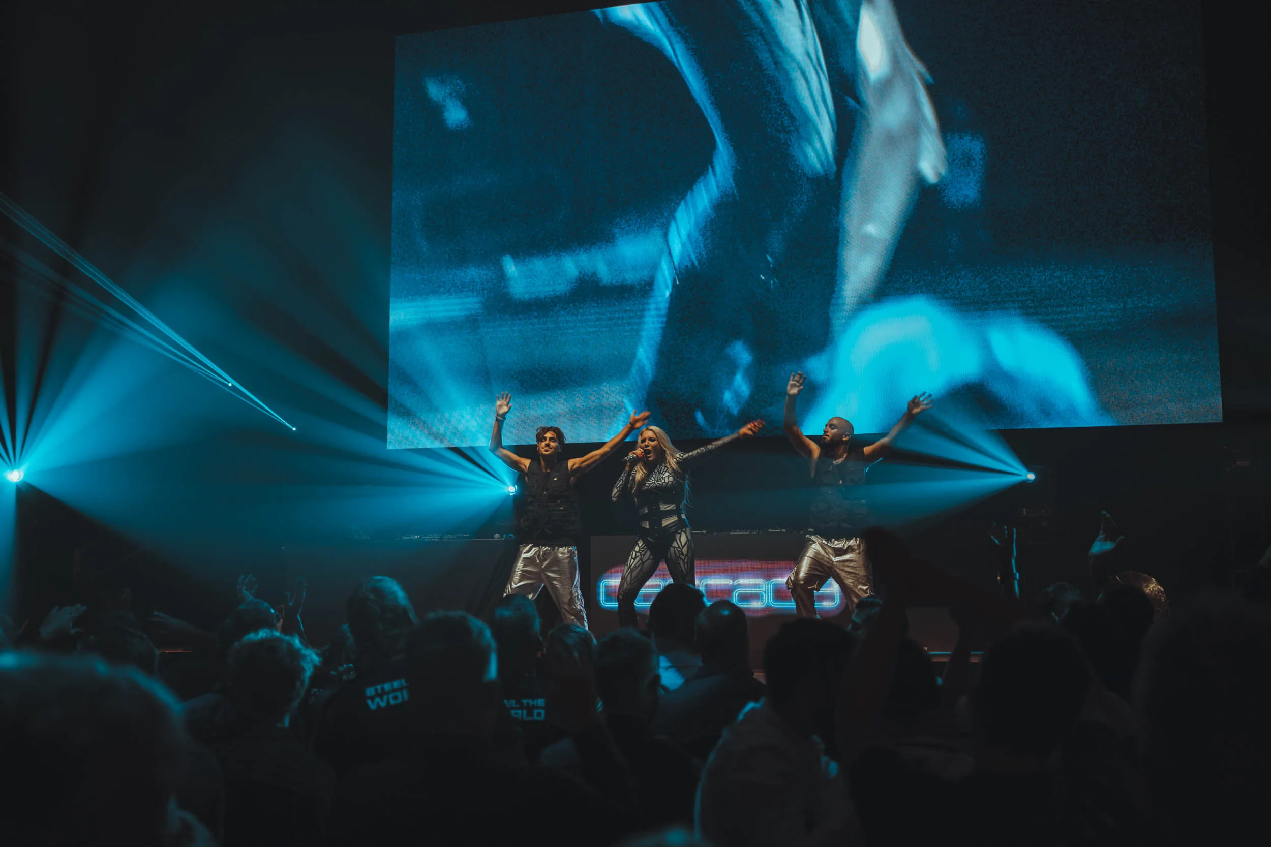 Dynamische Live-Performance mit drei Tänzern auf einer Bühne, spektakuläre Lichtshow in Blau, große LED-Leinwand im Hintergrund, energetisches Publikum beim Event.