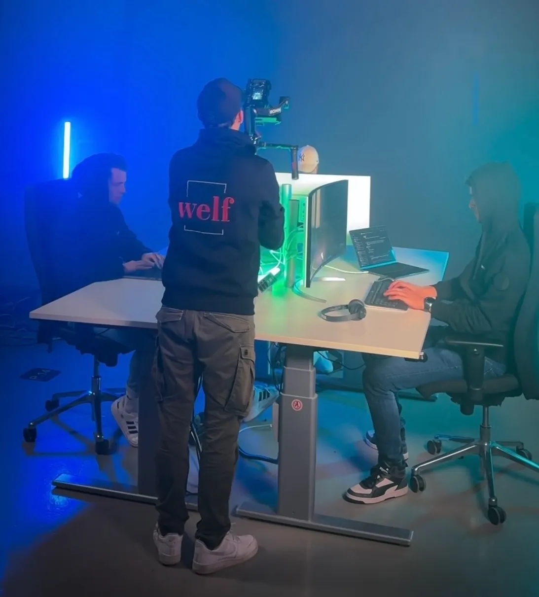 Behind-the-Scenes: Kameramann mit Gimbal filmt zwei Mitarbeitende am Schreibtisch, Studio-Setup mit blau-grünem Licht.