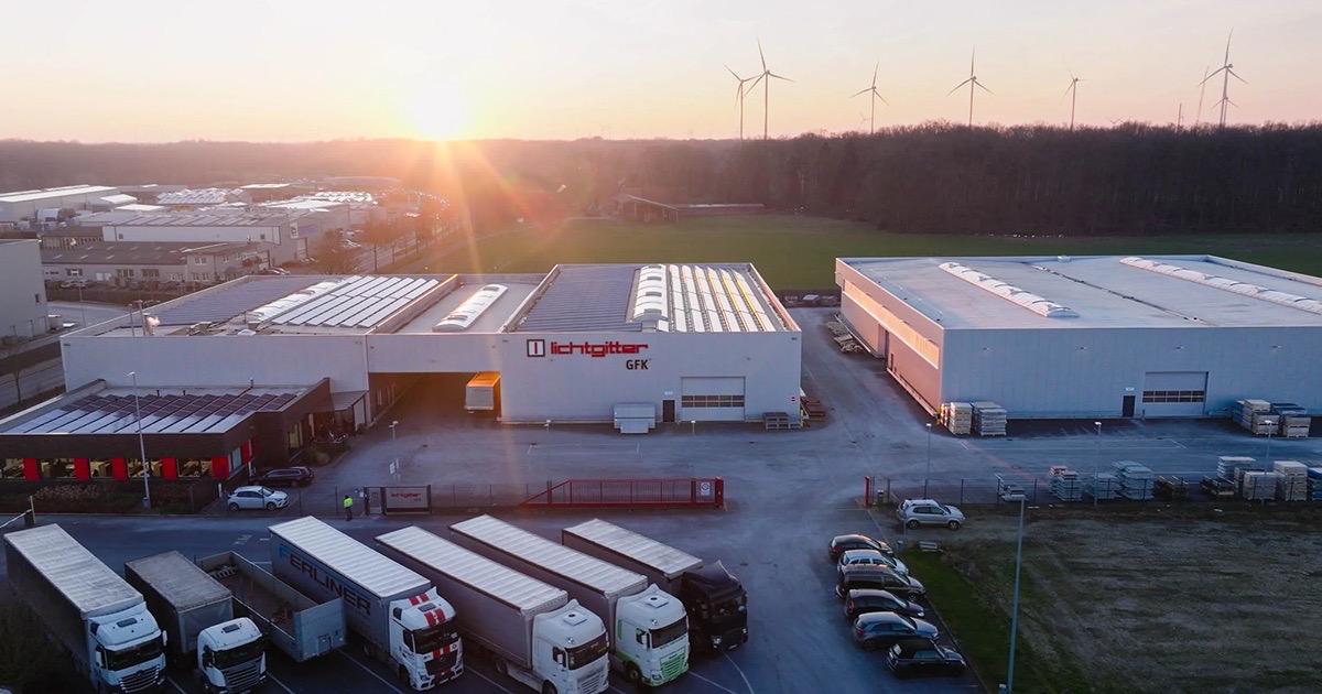 Drohnenaufnahme: Lichtgitter GFK Werk mit Lagerhallen und Lkw-Verkehr bei Sonnenuntergang, Windräder im Hintergrund.