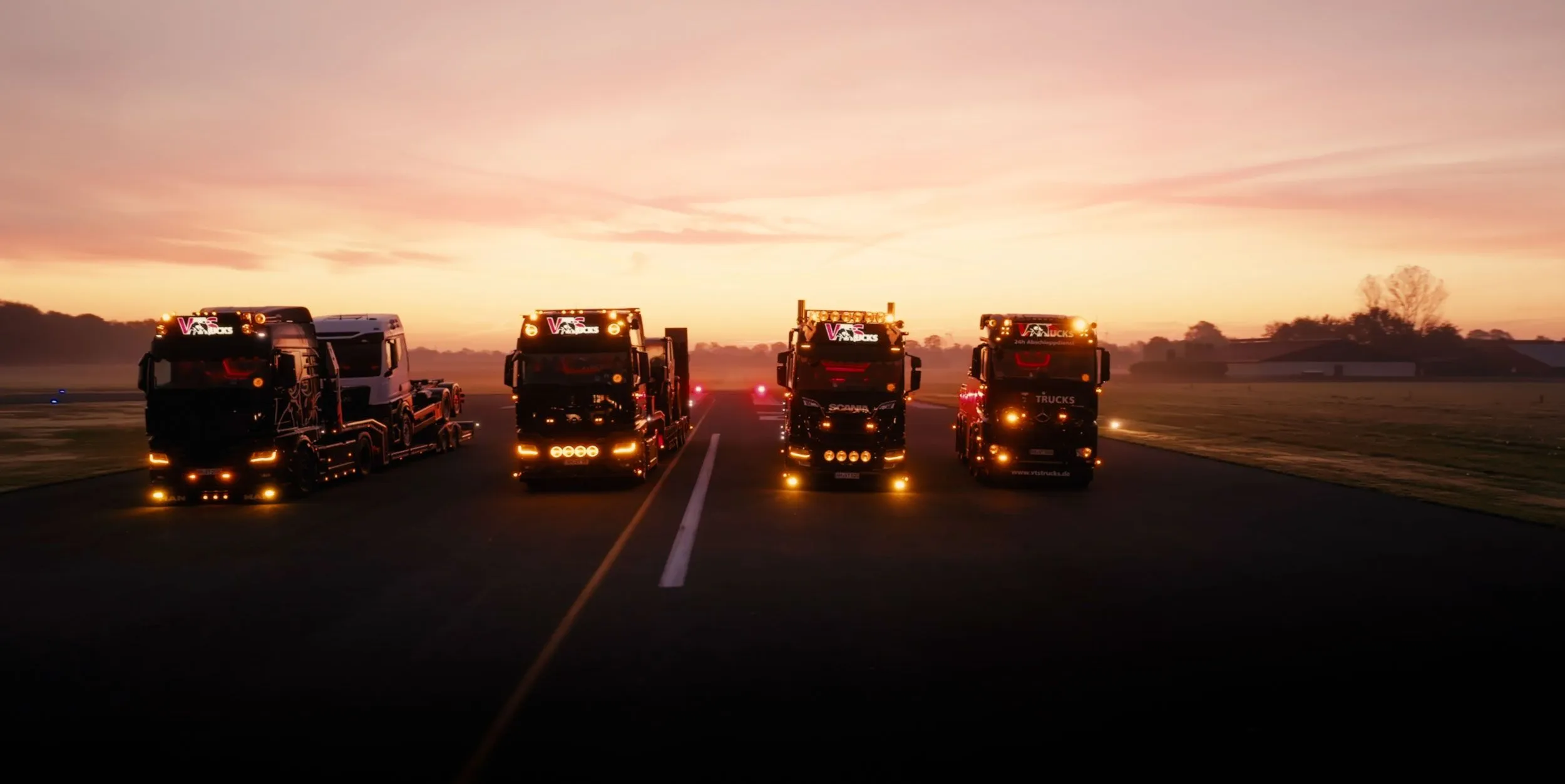 Vier beleuchtete Lkw nebeneinander auf gerader Strecke bei Sonnenaufgang.