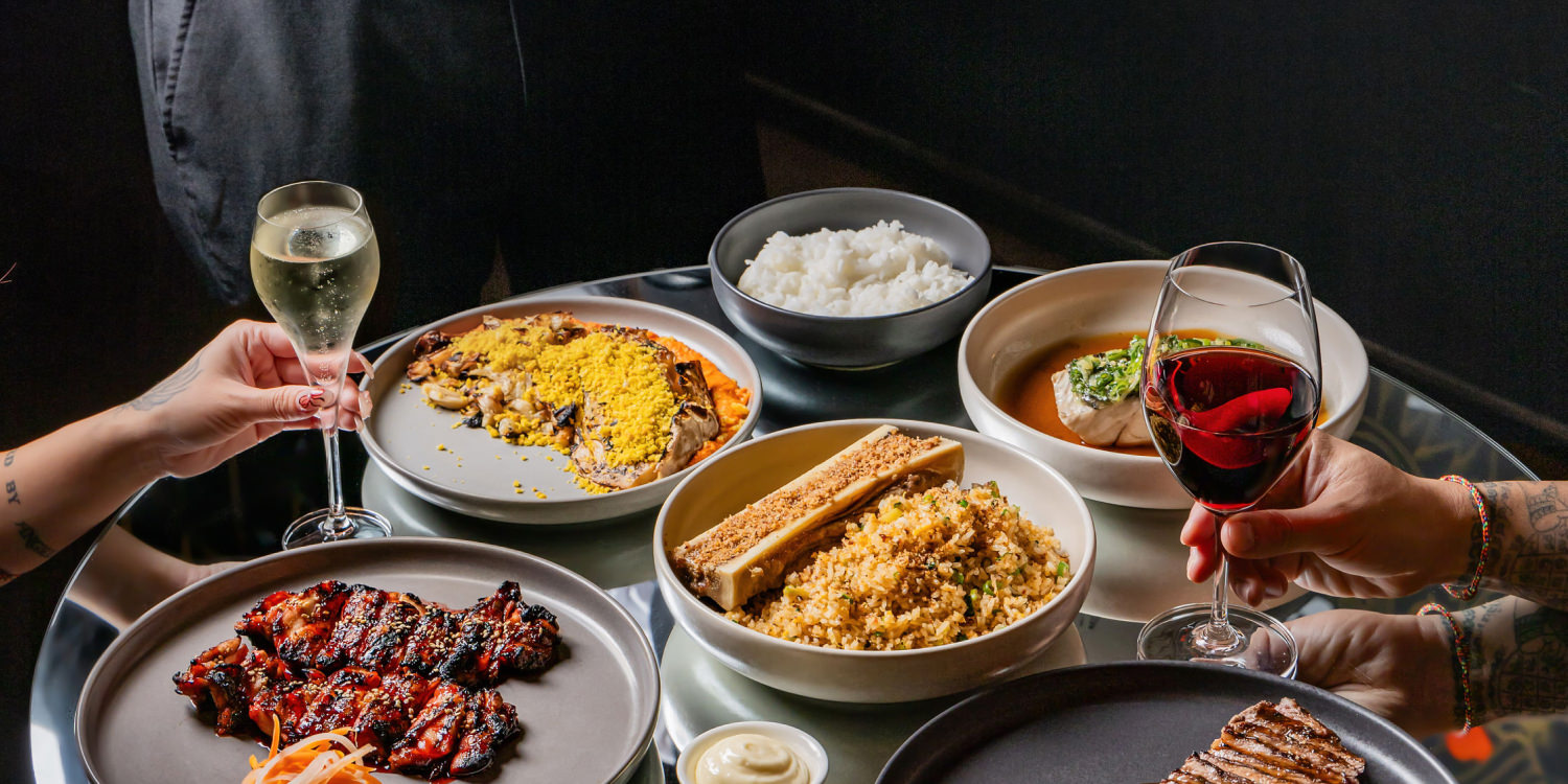 Table with various gourmet dishes including grilled meat, bone marrow with fried rice, a bowl of white rice, and a dish topped with yellow crumbs, accompanied by glasses of white and red wine held by tattooed hands.