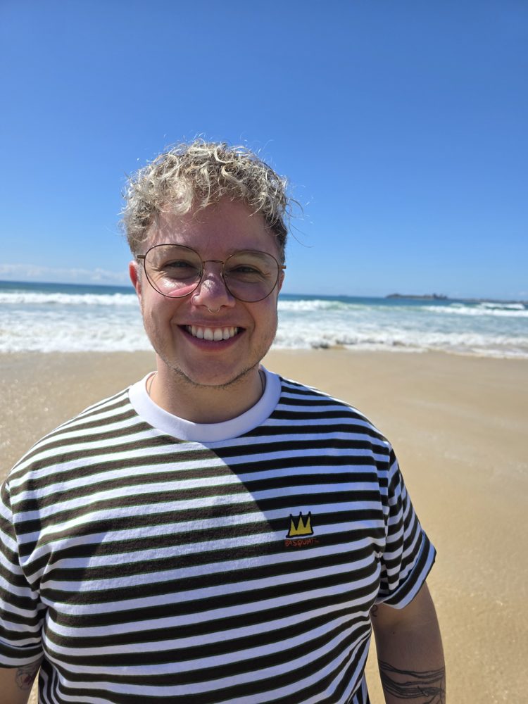 Leroy Maronian smiling in glasses and a black and white striped top on a sunny beach, with the ocean behind.