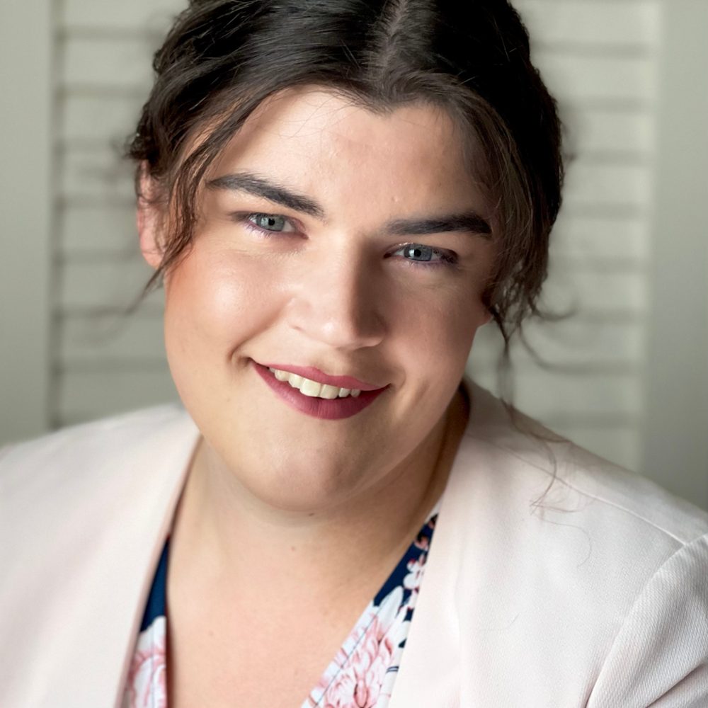 Maddie Sumner smiling, with dark hair pulled back and a pale pink jacket, in soft indoor light.