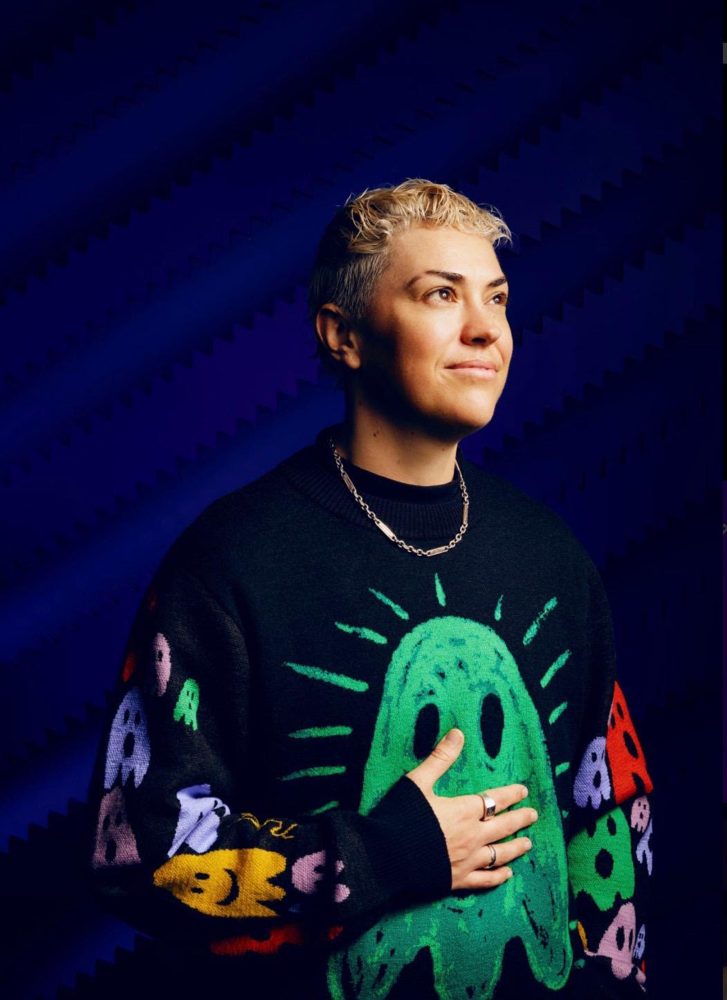 Harry Lloyd with short bleached hair, a silver chain and a black jumper with a green ghost design, looking off to the side against a dark blue backdrop.