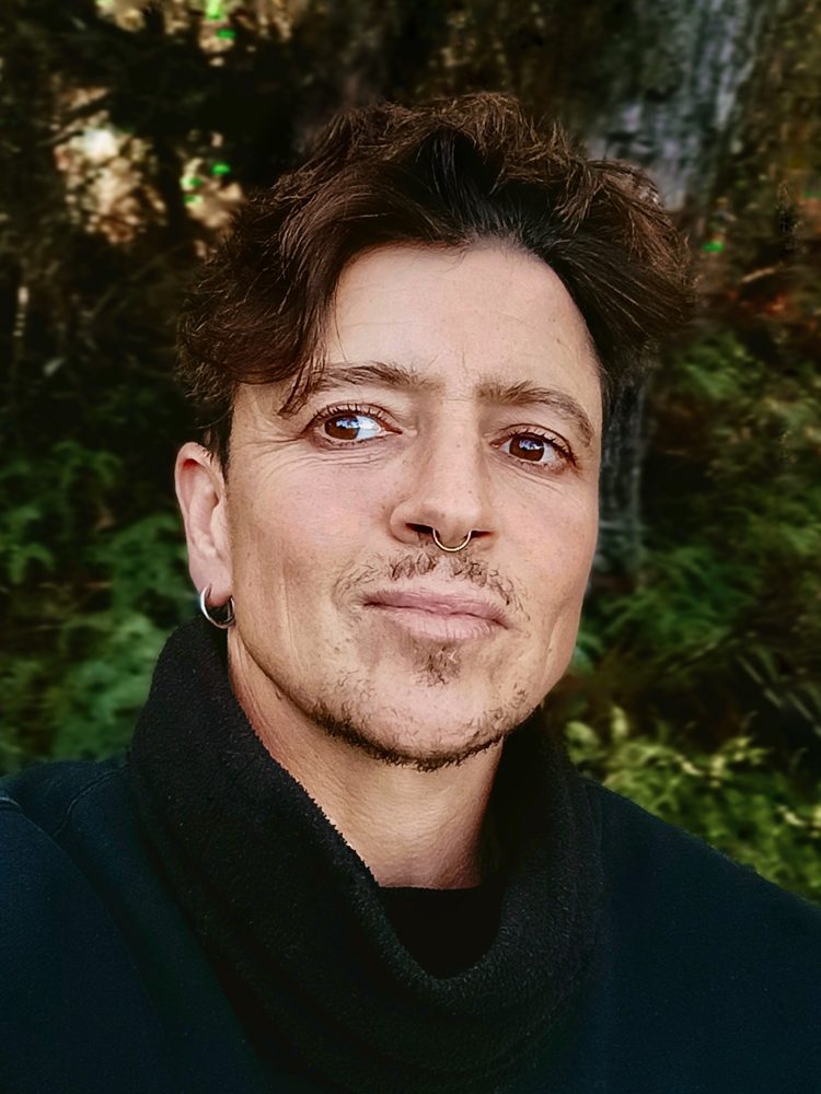 Close-up photo of Noa Wolf Smith Fletcher with tousled dark hair, a black scarf and a gentle smile, in front of green foliage.