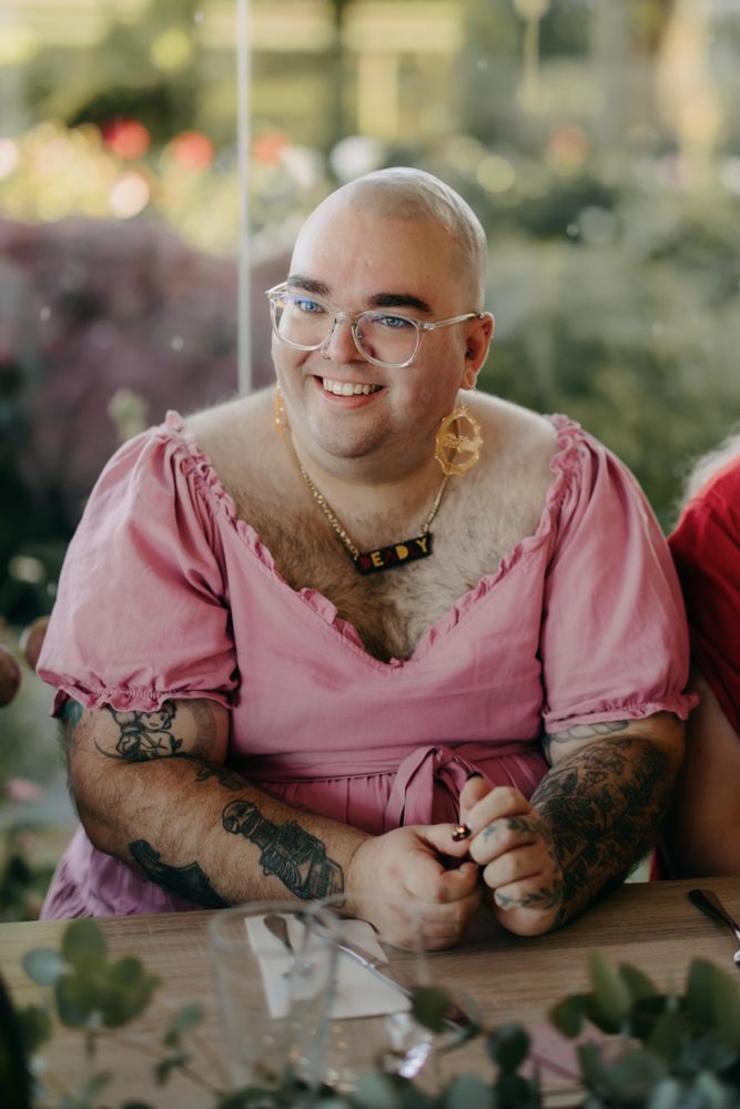 Nic Radoll smiling at an outdoor table in a pink ruffled dress, glasses and gold earrings, with a garden behind.