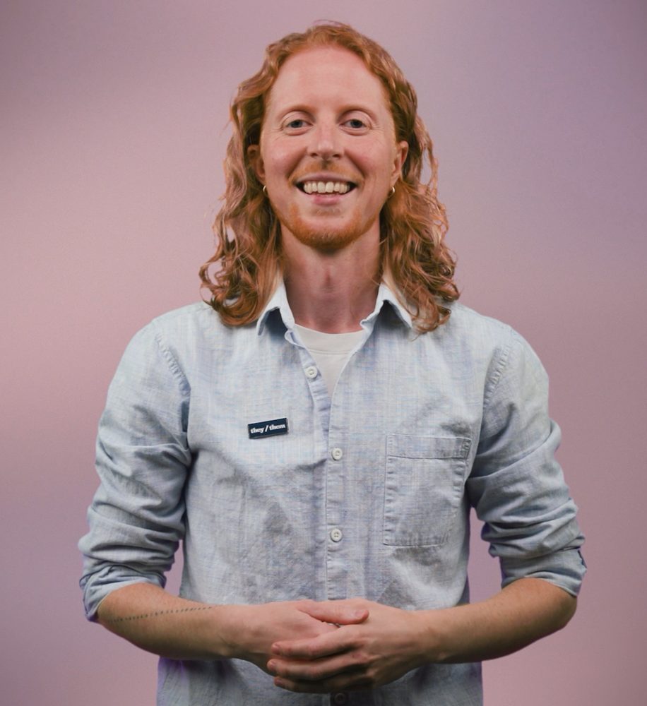 Ari Heart smiling, with long curly red hair and a light blue button-up shirt, hands clasped, against a pink backdrop.