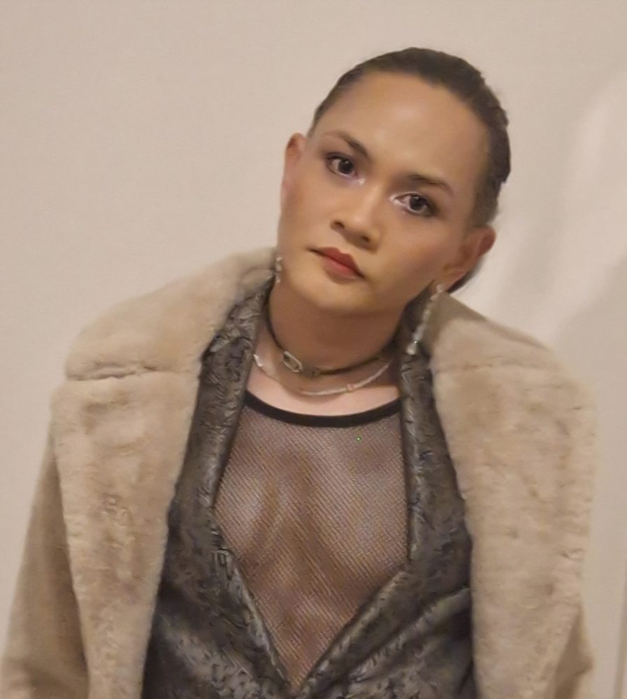 Budi Sudarto with hair pulled back, a faux fur coat over a mesh top and a calm expression, against a pale wall.