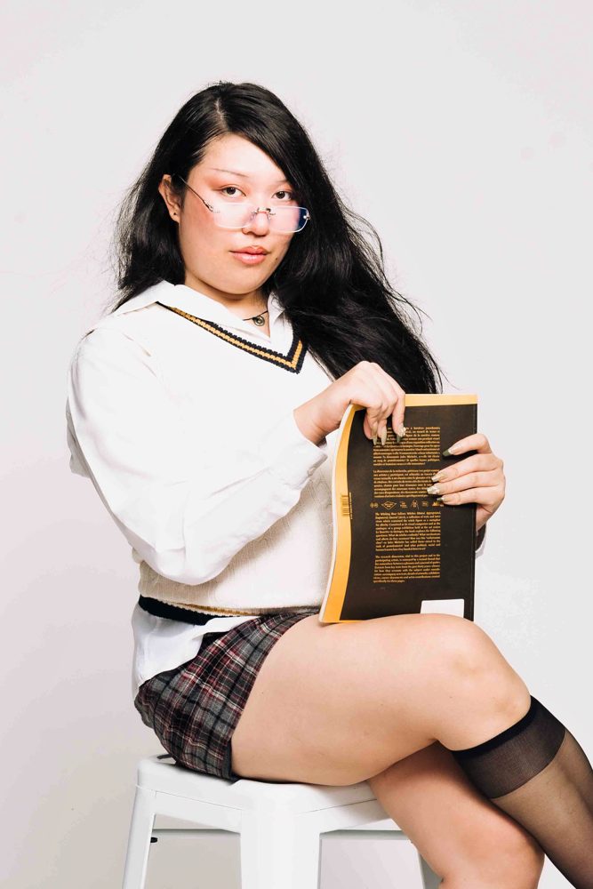 Aria Villero seated on a white stool holding a book, wearing glasses, a white V-neck jumper and a plaid skirt, against a white backdrop.