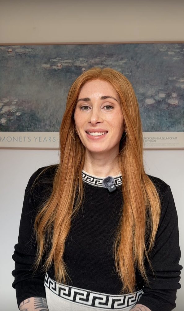 Leta Taylor smiling, with long red hair and a black top, standing in front of a framed artwork.