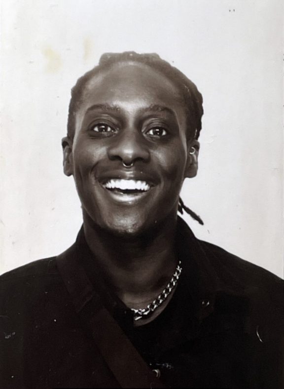 Black and white photo of Shei Udeagu with a wide smile, braided hair, a chain necklace and a dark top.