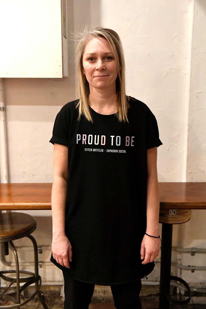 T Edgell-Hall standing in a room with wooden stools, with long blonde hair and a black T-shirt reading Proud to Be.