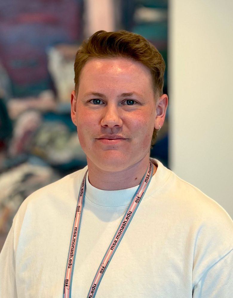 Connor Bayly-Jones with short hair, a white top and a lanyard and a gentle smile, indoors.