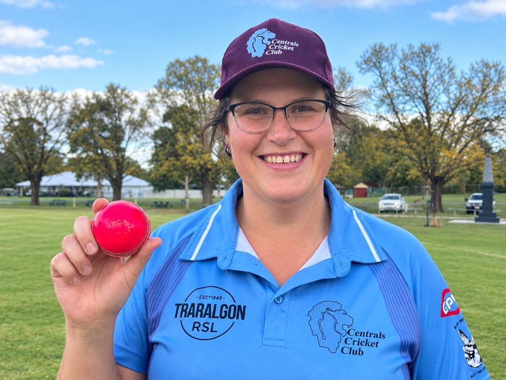 Kylie Collins smiling on a sports oval in a blue Centrals Cricket Club shirt and cap, holding up a pink cricket ball.