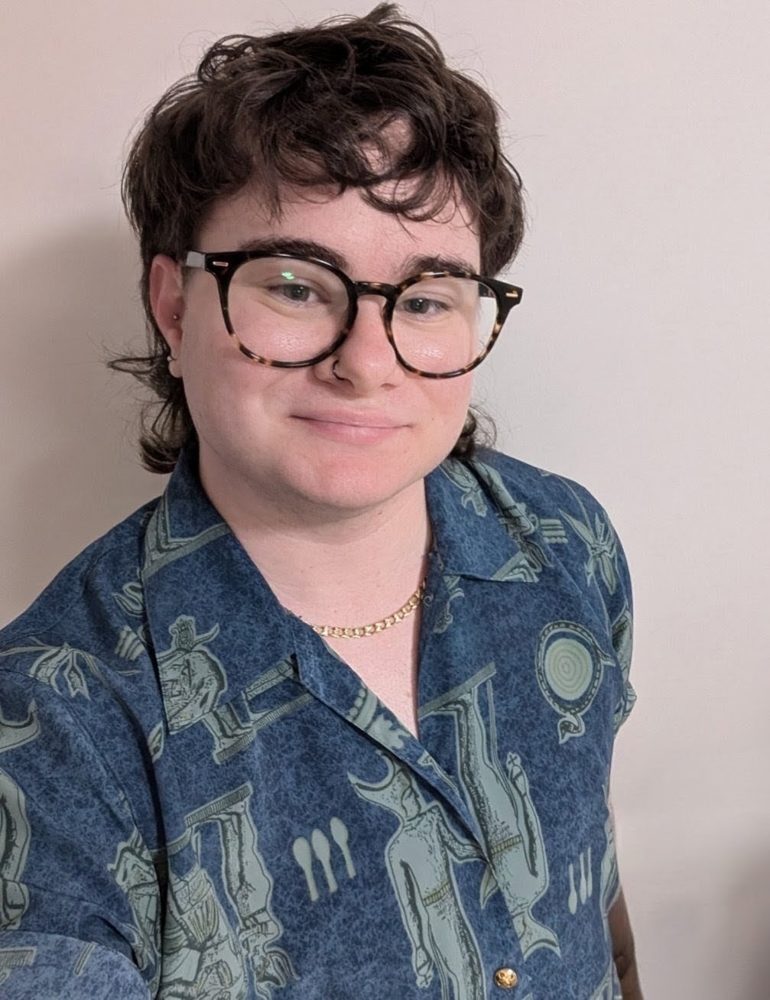 Luka Ritter with short dark curly hair, glasses, a patterned blue button-up shirt and a small smile, indoors.
