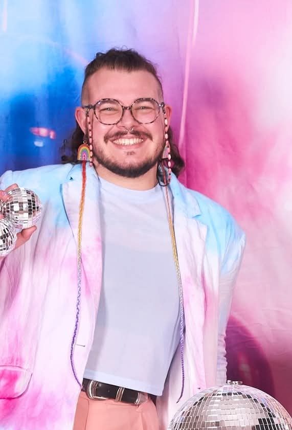 James Bush beaming in glasses, a pastel jacket and white T-shirt, holding disco balls in front of a pink and blue backdrop.