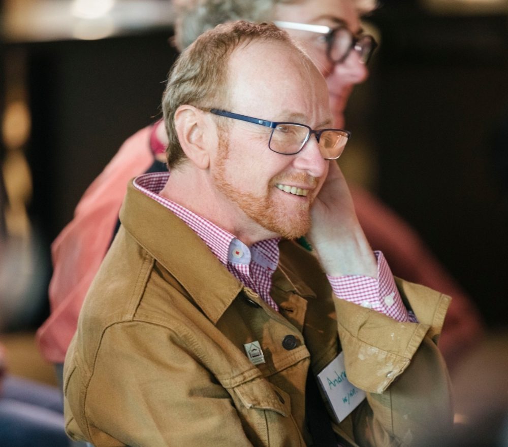 Andrew Eklund smiling in glasses and a tan jacket over a checked shirt, at a community event.