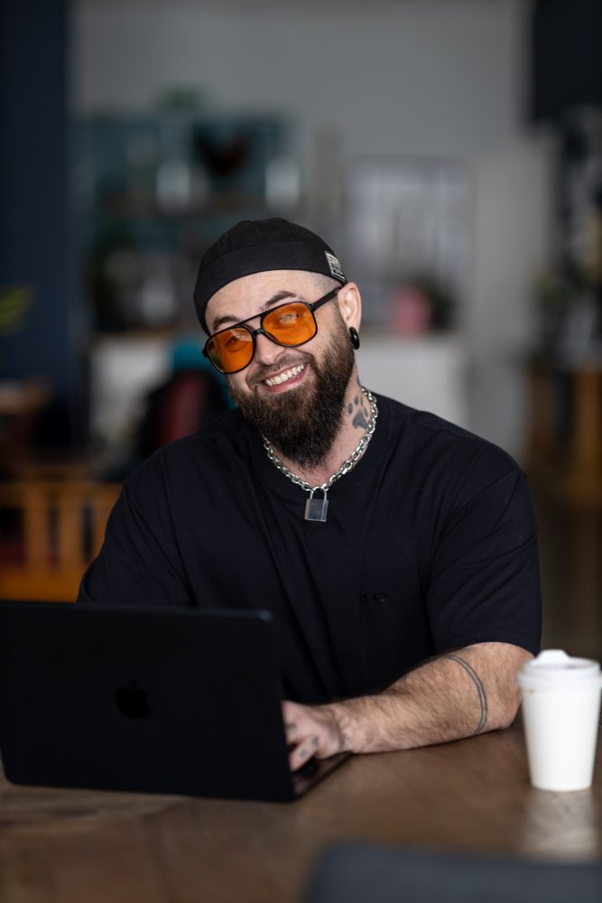 Sam Dengate smiling at a café table with a laptop and a coffee cup, wearing orange-tinted glasses, a black cap and a black T-shirt.