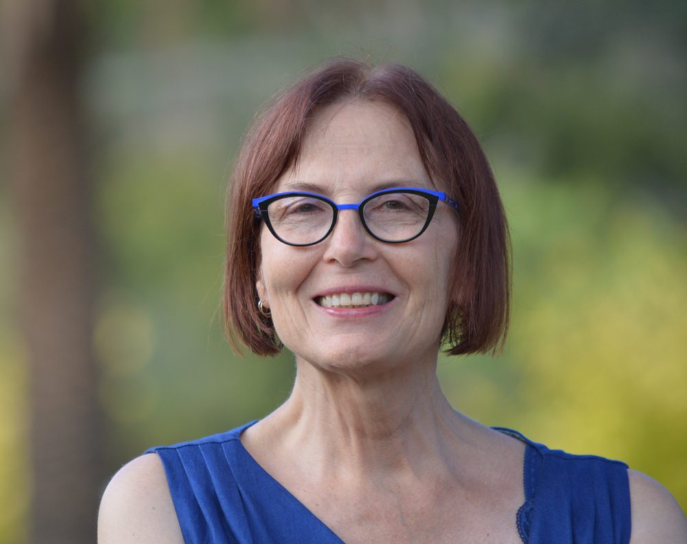 Julie Peters smiling, with short auburn hair, blue glasses and a blue top, against leafy greenery.