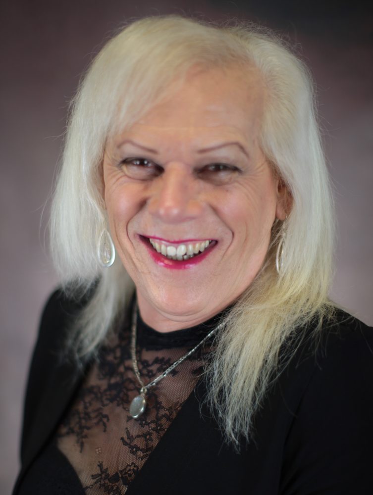 Greer McGearey with a wide smile, long platinum hair, a black lace top and a pendant necklace, against a grey backdrop.