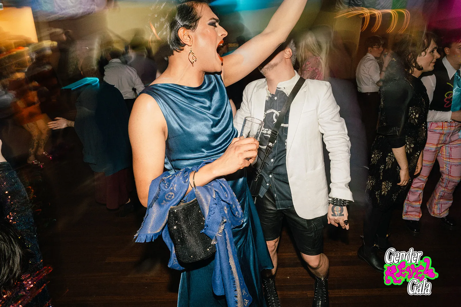 A guest in a blue satin gown dancing with one arm raised on a busy dance floor at the Gender Revel Gala