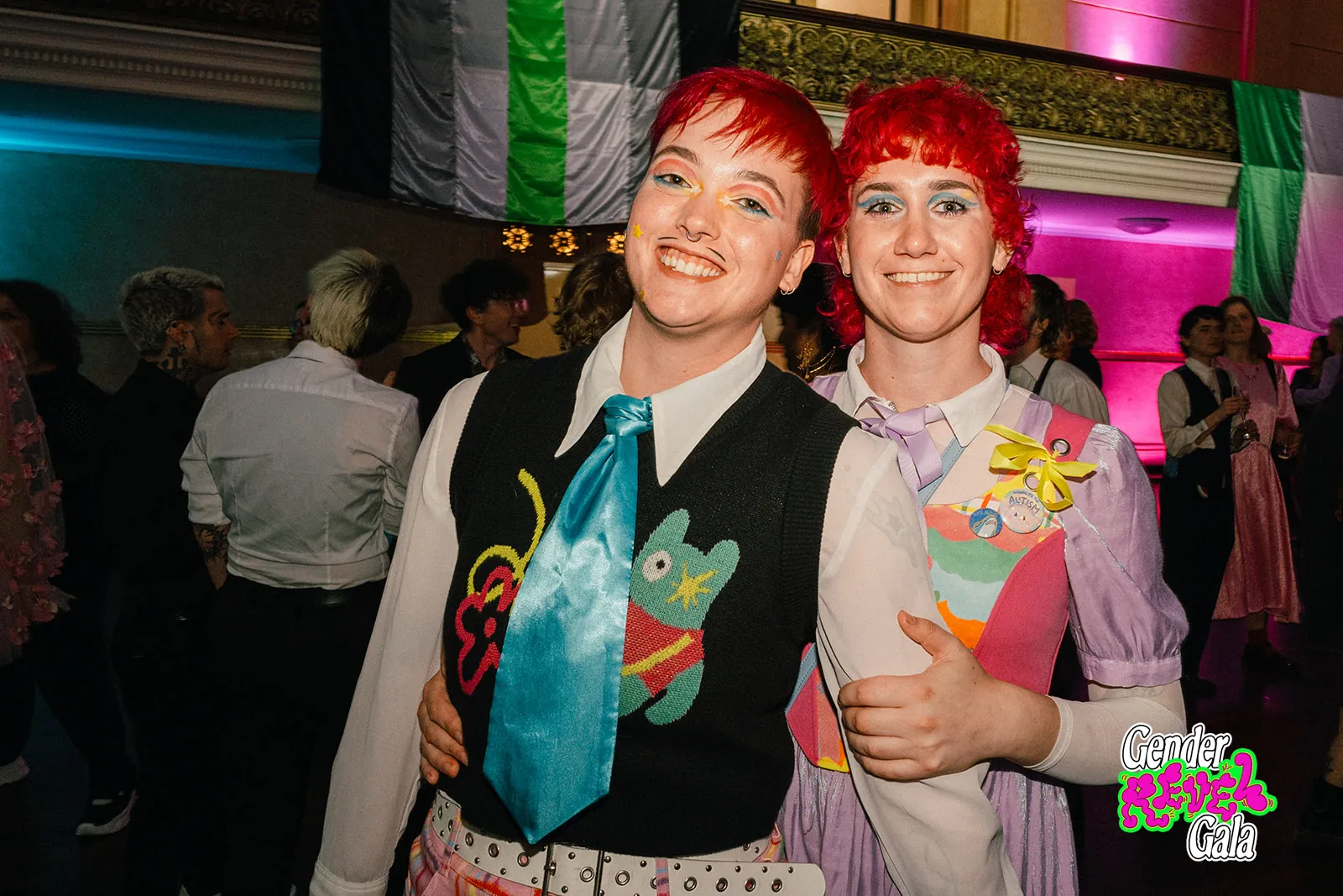 Two guests with bright red hair and colourful outfits smiling together under trans flags at the Gender Revel Gala