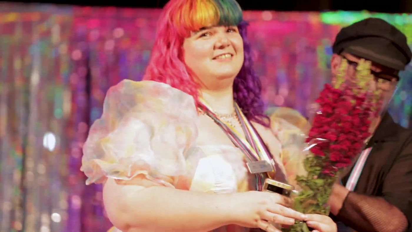 A guest with rainbow hair and a frilly white dress holding red flowers