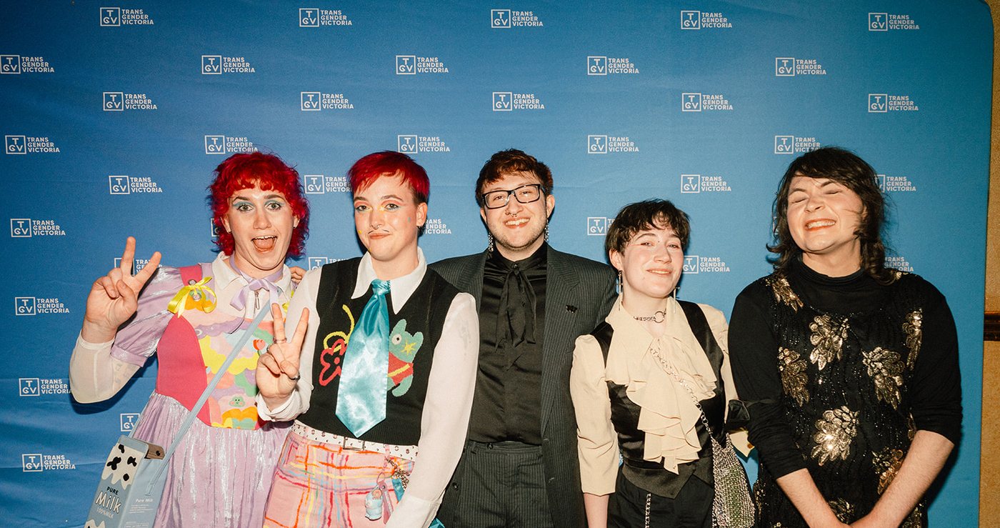 Five guests in colourful outfits posing in front of the Transgender Victoria media wall at the Gala