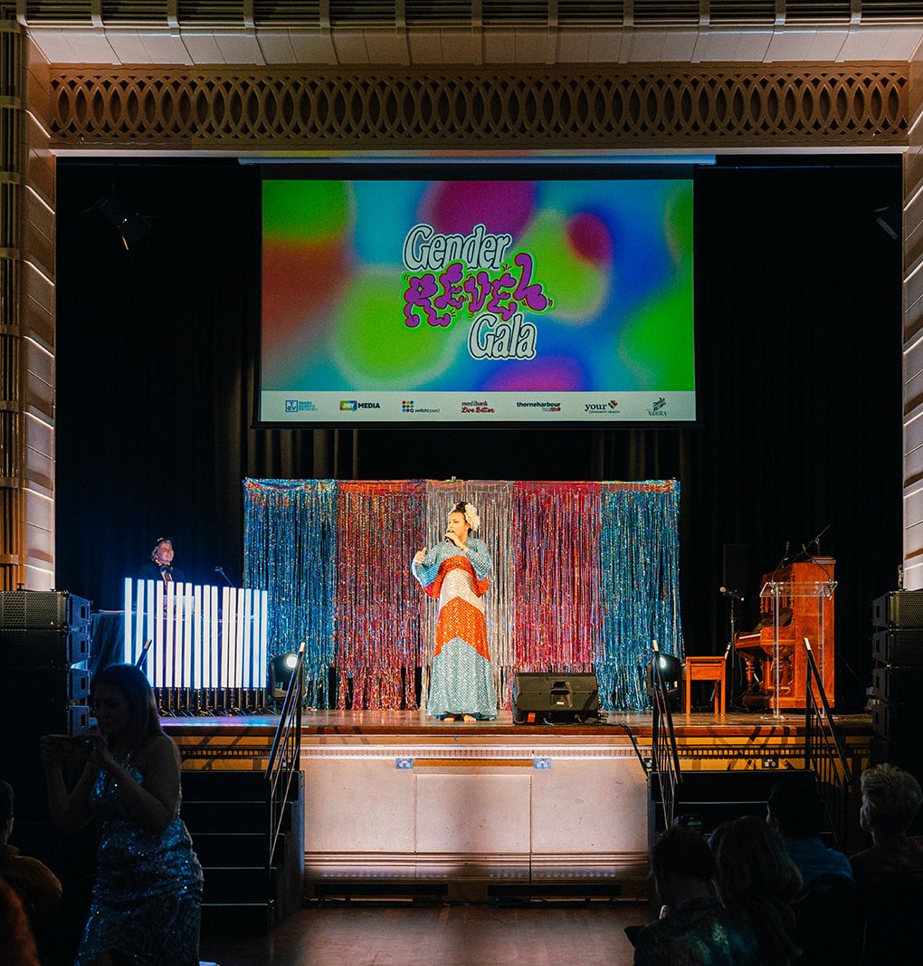 The Gala stage with a performer in front of a tinsel curtain and the Gender Revel Gala logo on the big screen