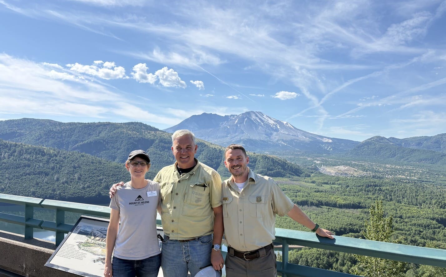 Three people posing on an overlook with Mount St. Helens and forested hills in the background.