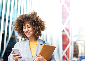 A person with curly hair smiling down at their smartphone and holding a tan notebook.