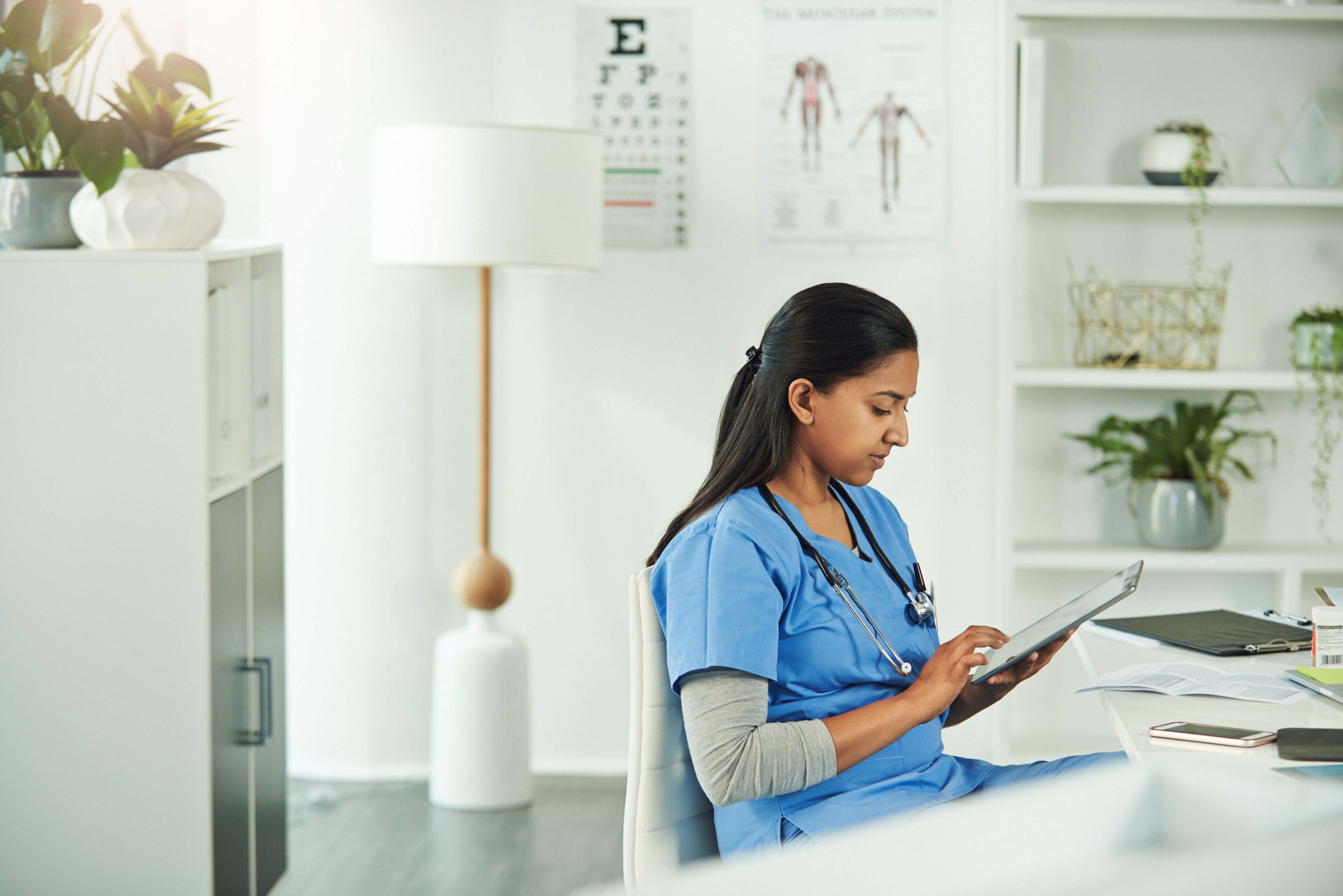 ​​A medical professional in blue scrubs and a stethoscope focused on a tablet at a desk in a well-organized office with health posters and plants.