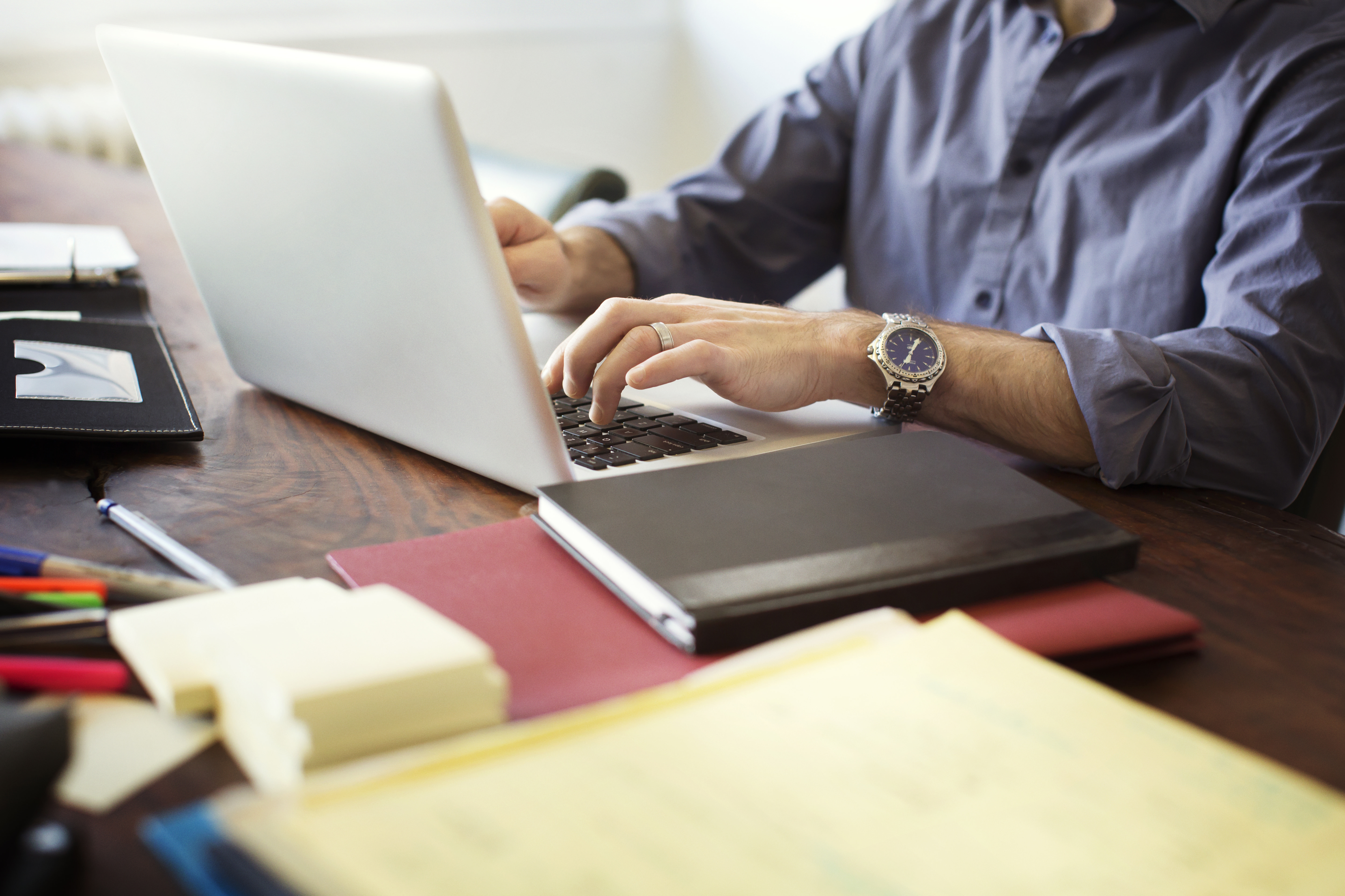 Osoba pracująca na laptopie przy biurku z notatnikami i zegarkiem na nadgarstku.