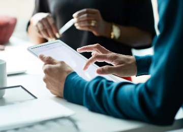 Hands using a tablet on a desk with another person in the background.