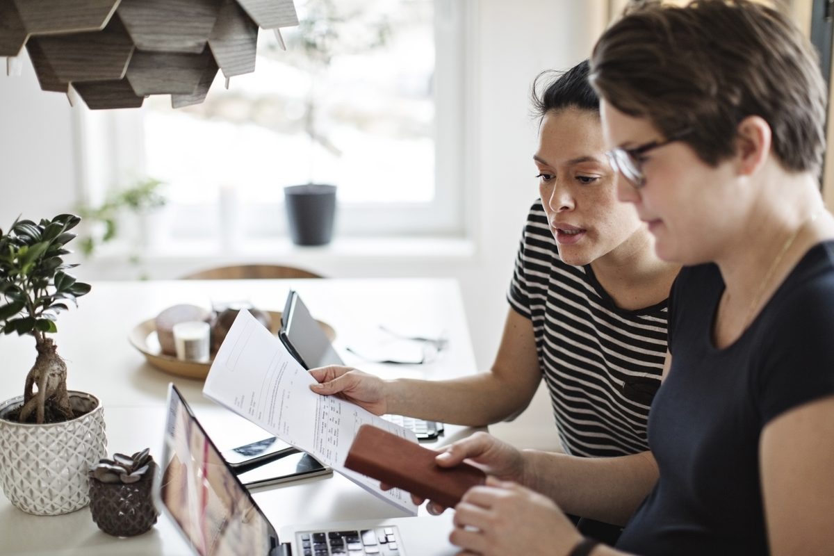 Two people at a kitchen table look over a printed document, with a laptop open beside them.