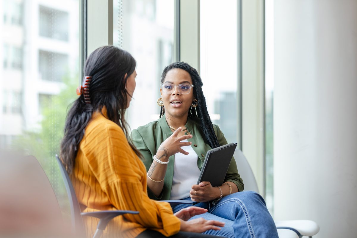 Two people talk by a window, one holding a tablet.