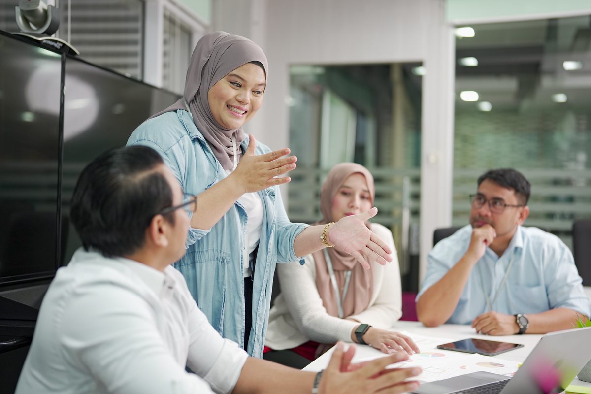 A woman standing at a conference table gestures while three colleagues listen, with charts and a laptop on the table.