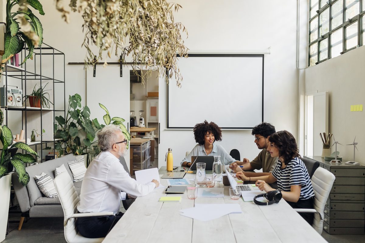 Four people sit around a long table in a bright office, with laptops and papers spread out in front of them.
