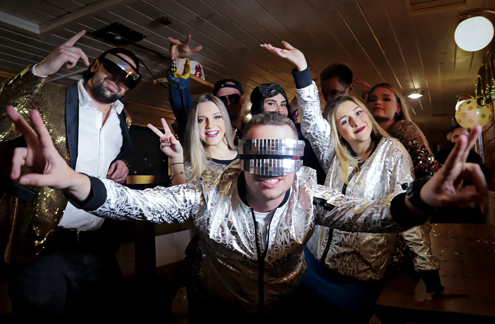 Skupina mladých lidí v lesklém oblečení tančících a pózujících na večírku v interiéru.