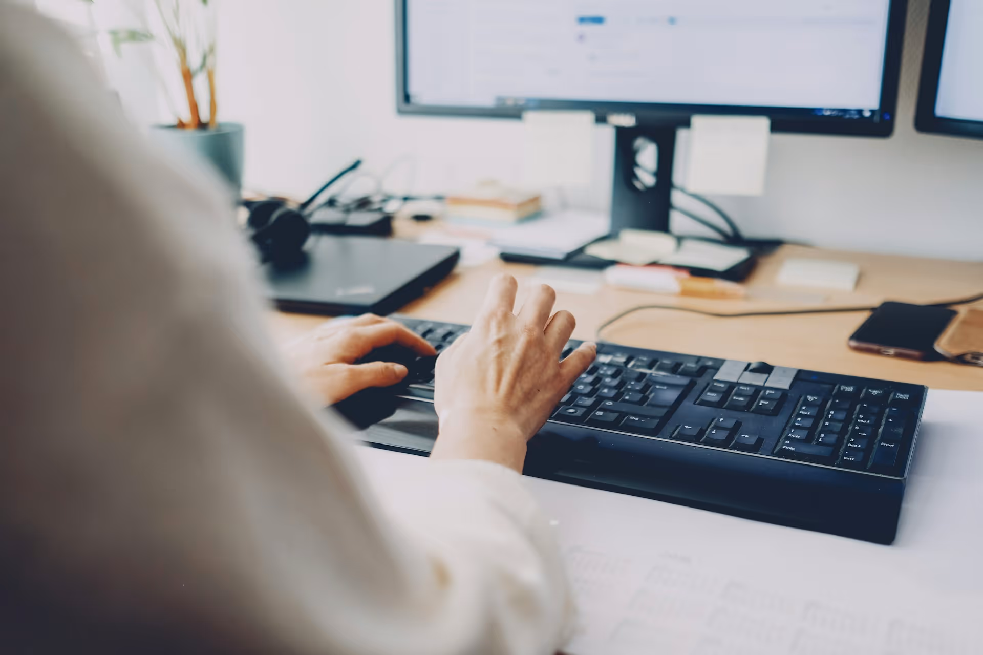 Person typing on a black keyboard at a desk with dual monitors and office supplies.