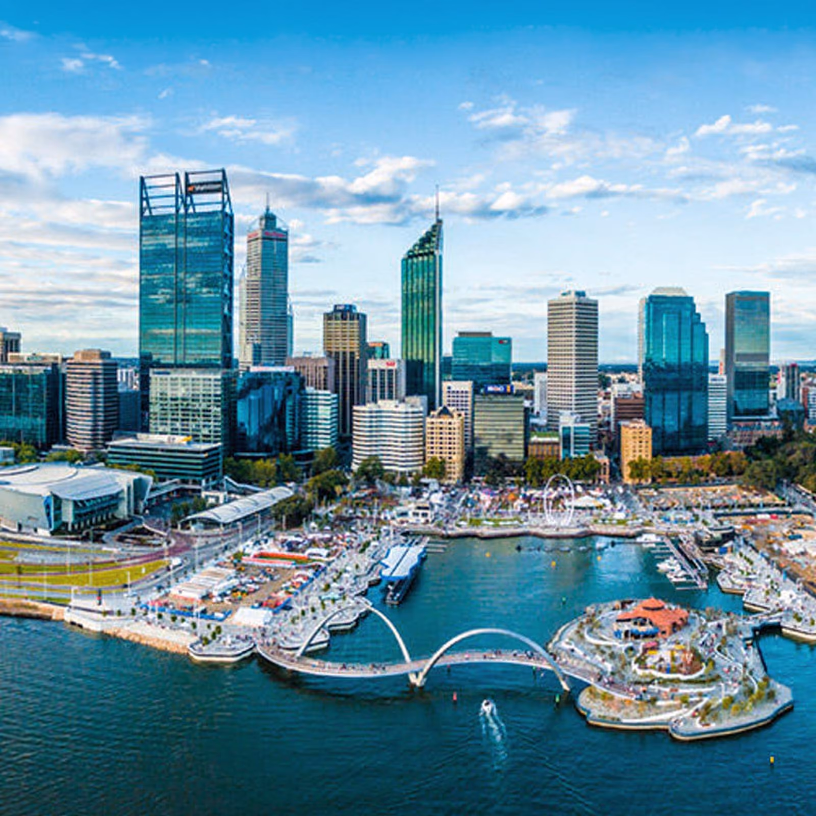 Aerial view of a Perth city skyline with modern high-rise buildings, a curved pedestrian bridge over the water, boats, and a partly cloudy sky.